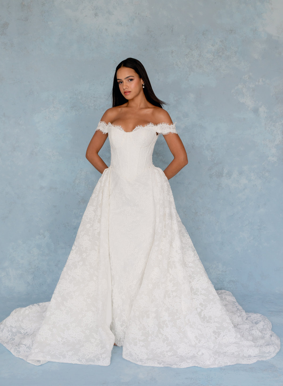 A woman modeling an off-the-shoulder white lace wedding gown with a fitted bodice and full skirt, standing against a textured light blue backdrop. / All over lace column gown with a dramatic matching overskirt.
