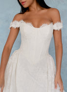 An off-the-shoulder white lace dress with a fitted corset-style bodice and a full skirt, shown on a model against a textured light blue backdrop. / All over lace column gown with a dramatic matching overskirt.