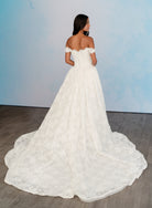An off-the-shoulder white lace wedding gown with a full skirt and long train, shown from the back on a model against a light blue studio backdrop. / All over lace column gown with a dramatic matching overskirt.