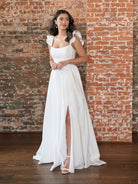 Woman in a white dress standing against a brick wall. / This clean, satin gown has encased boning on the bodice and a feminine, ruffle sleeve. The skirt has soft gathers and is finished at the waist with a self-tie belt.