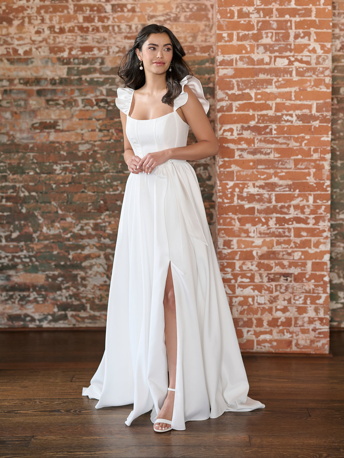 Woman in a white dress standing against a brick wall. / This clean, satin gown has encased boning on the bodice and a feminine, ruffle sleeve. The skirt has soft gathers and is finished at the waist with a self-tie belt.