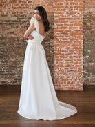 Woman in a white wedding dress standing against a brick wall. / This clean, satin gown has encased boning on the bodice and a feminine, ruffle sleeve. The skirt has soft gathers and is finished at the waist with a self-tie belt.