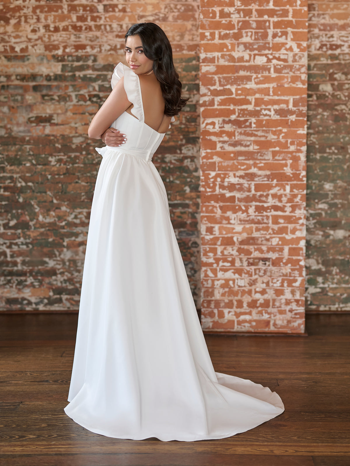 Woman in a white wedding dress standing against a brick wall. / This clean, satin gown has encased boning on the bodice and a feminine, ruffle sleeve. The skirt has soft gathers and is finished at the waist with a self-tie belt.