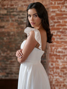 A woman wearing a white sleeveless dress with ruffled shoulder details, standing in front of a rustic red brick wall. / This clean, satin gown has encased boning on the bodice and a feminine, ruffle sleeve. The skirt has soft gathers and is finished at the waist with a self-tie belt.
