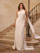 Woman in a white halter neck gown standing on a staircase with stone walls. / Halter stretch crepe trumpet with self scarf.