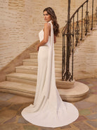 Woman in a white gown standing on a staircase with stone walls. / Halter stretch crepe trumpet with self scarf.