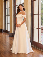 Woman in a white off-shoulder gown standing in a room with large windows. / Draped off the shoulder satin A-line.