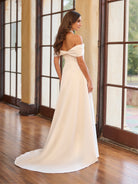 Woman in a white off-shoulder gown standing in a room with large windows. / Draped off the shoulder satin A-line.