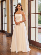 Woman in a white strapless gown standing indoors with large windows in the background / Draped off the shoulder satin A-line.