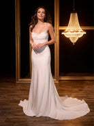 Woman in a white evening gown standing in a room with a chandelier. / Slim gown with cowl neckline and low, dramatic back detail.