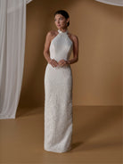 A woman modeling a sleeveless, high-neck, floor-length white dress with a smooth fitted bodice and textured lace overlay from the knees down, posed against a warm tan studio backdrop with draped white fabric on the left. / With lined skirt. Column gown with halter neckline and lace detailing.