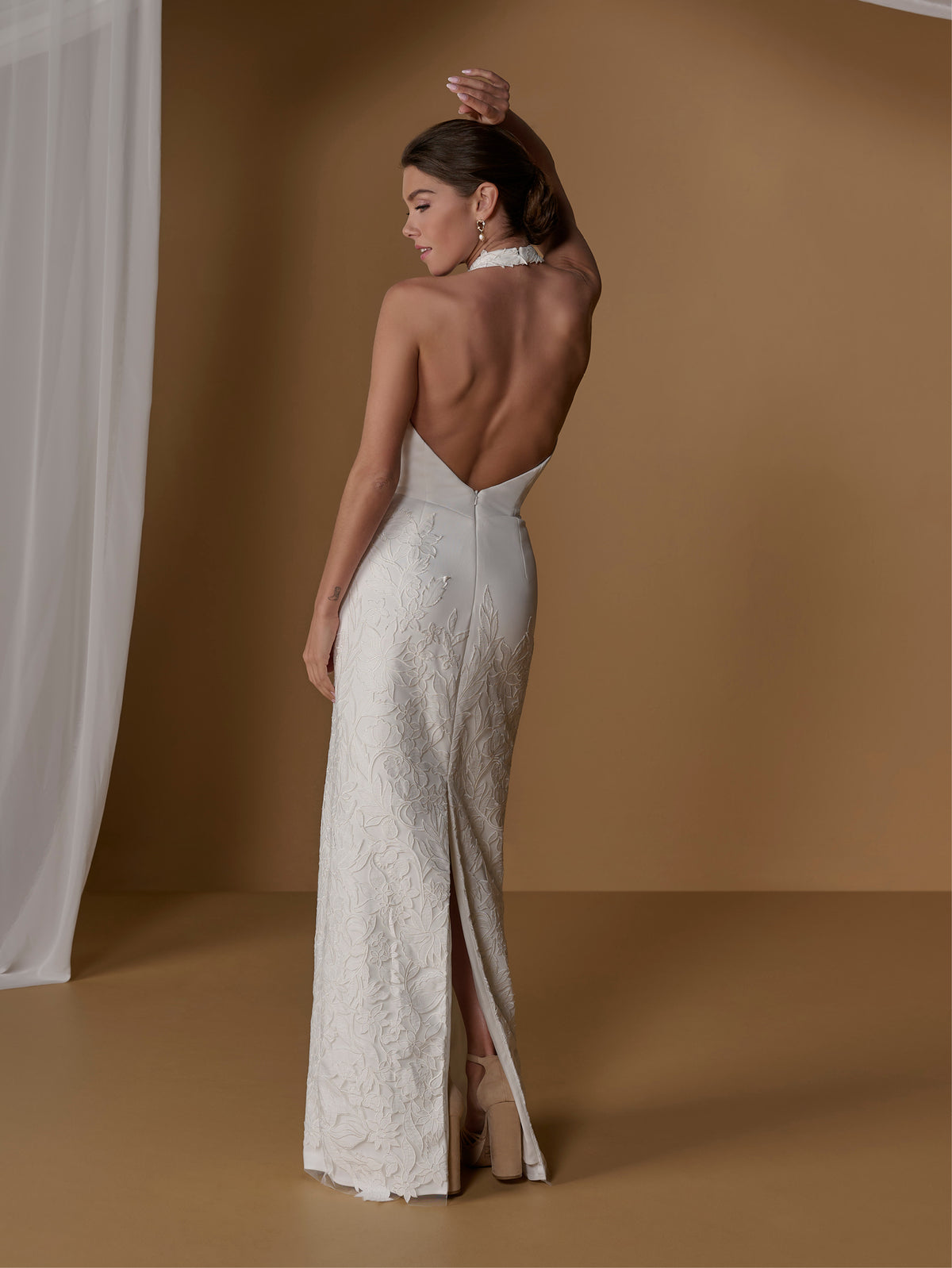 A woman modeling a sleeveless, backless ivory lace gown with a high slit, shown from the back against a warm tan studio backdrop with a white curtain on the left. / With lined skirt. Column gown with halter neckline and lace detailing.