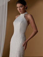 A woman modeling a fitted sleeveless wedding dress in warm off-white/ivory with floral lace applique at the bodice and along the skirt, posed against a tan studio backdrop with a white curtain at the side. / With lined skirt. Column gown with halter neckline and lace detailing.