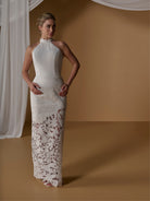 A woman models a sleeveless, high-neck floor-length wedding gown with a fitted bodice and intricate floral lace overlay on the skirt, posed against a warm tan studio backdrop with draped white fabric. / Unlined skirt. Column gown with halter neckline and lace detailing.