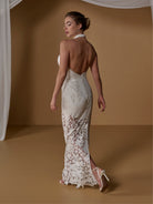 A woman modeling a sleeveless, open-back halter-neck wedding gown with intricate lace detailing, shown from behind against a warm brown studio backdrop with draped white fabric. / Unlined skirt. Column gown with halter neckline and lace detailing.
