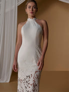 A woman modeling a sleeveless halter-neck white dress with floral lace applique at the high neckline and along the lower skirt, posed against a warm tan studio backdrop with draped white fabric on the left. / Unlined skirt. Column gown with halter neckline and lace detailing.