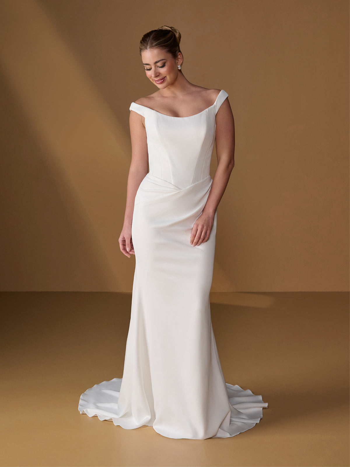 A woman modeling an off-the-shoulder white wedding gown with a fitted bodice, draped skirt, and a short train against a warm tan studio backdrop. / Tip of the shoulder gown with v waist and lace up back.