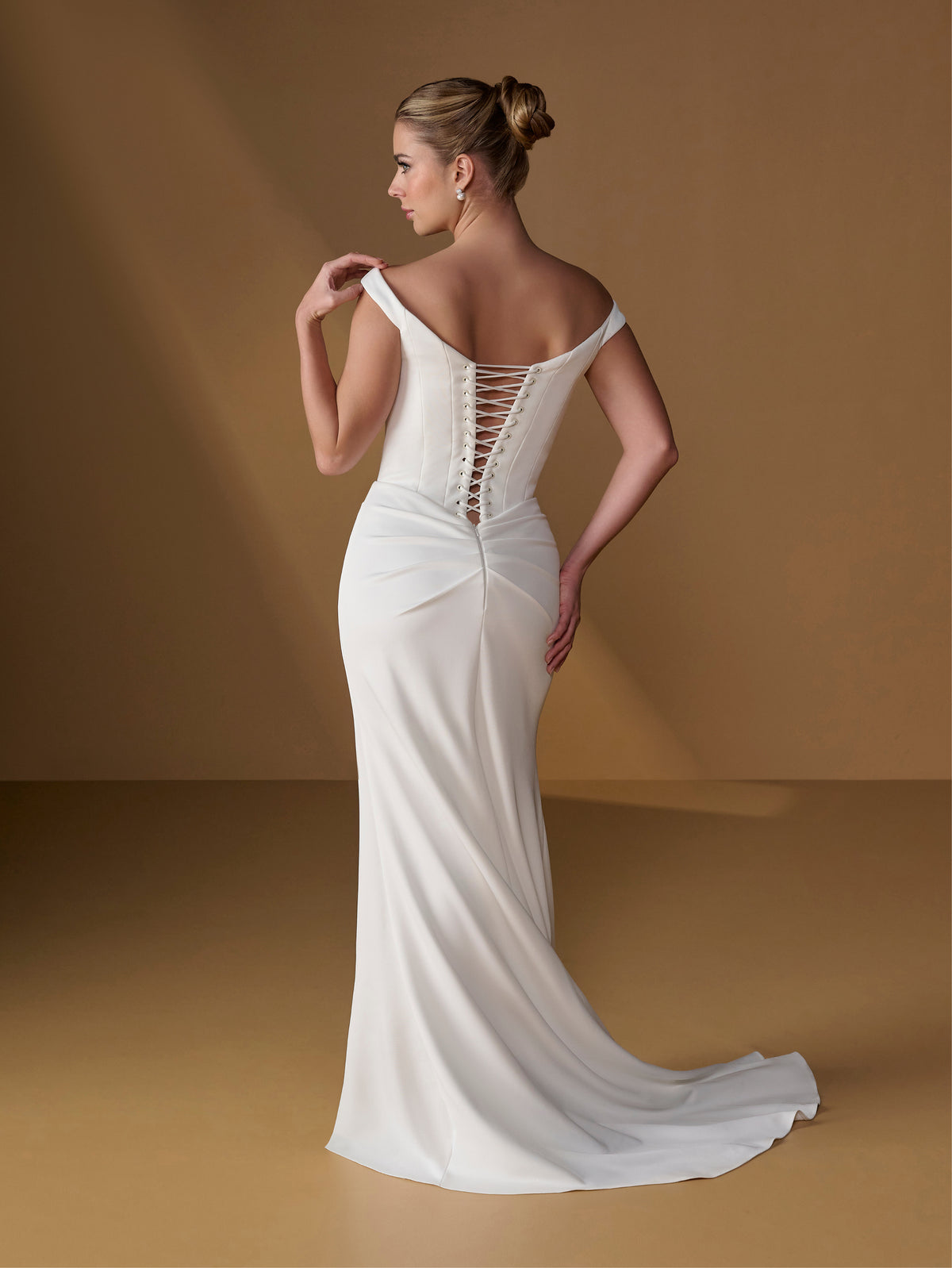 A woman modeling a fitted off-the-shoulder wedding gown from the back, showing a lace-up open back and long train, against a warm tan studio backdrop. / Tip of the shoulder gown with v waist and lace up back.