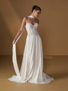 A woman models a strapless white wedding gown with a lace corset bodice, flowing chiffon skirt, and a long matching veil against a warm tan studio backdrop. / Faux 2-piece gown with lace bodice and full skirt.