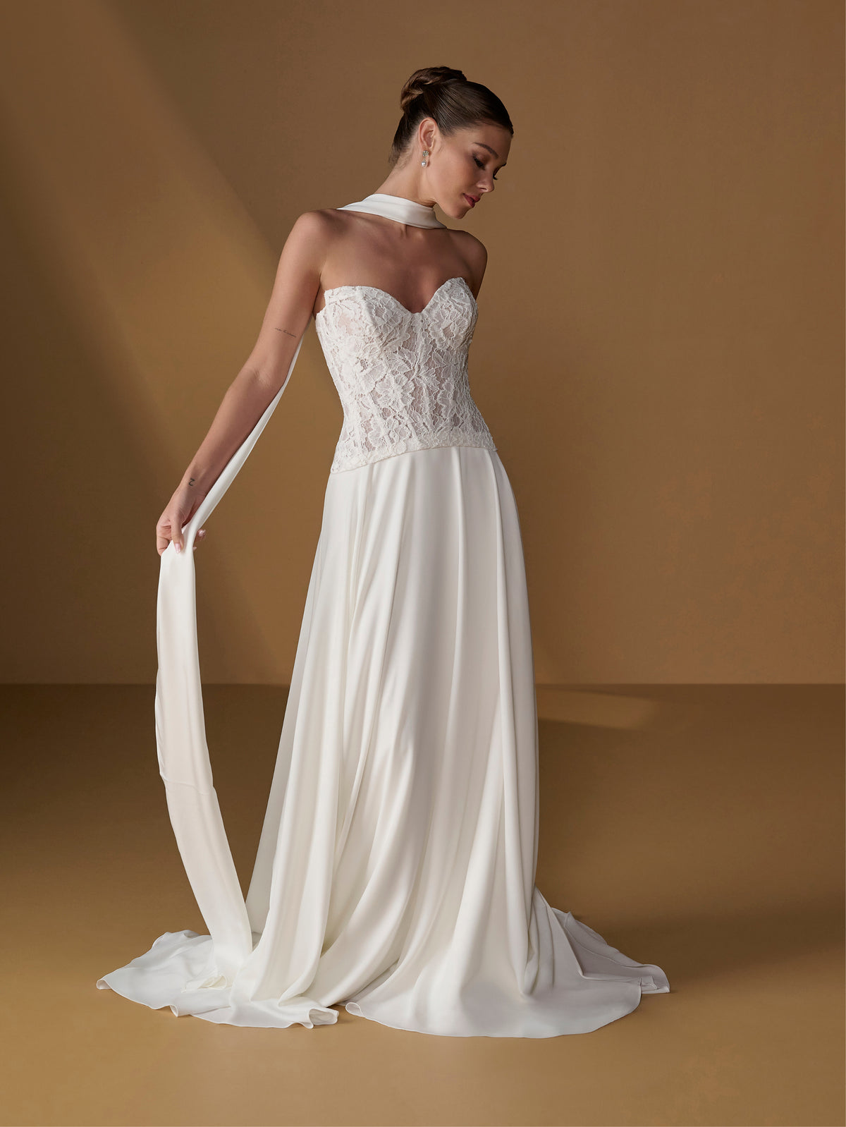 A woman models a strapless white wedding gown with a lace corset bodice, flowing chiffon skirt, and a long matching veil against a warm tan studio backdrop. / Faux 2-piece gown with lace bodice and full skirt.