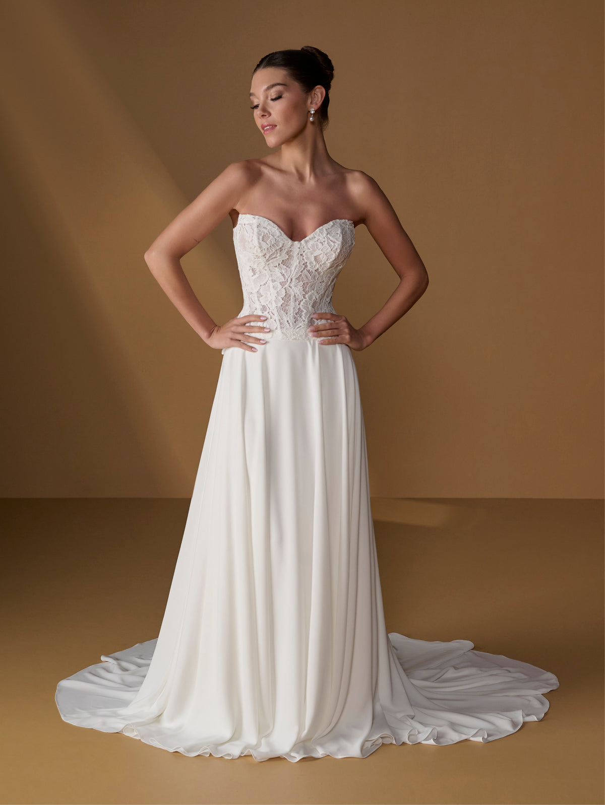 A woman modeling a strapless white wedding gown with a lace-embellished sweetheart bodice and flowing satin skirt with a train, posed against a warm tan studio backdrop. / Faux 2-piece gown with lace bodice and full skirt.