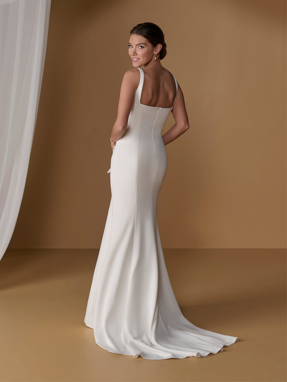 A woman modeling a sleeveless off-white mermaid-style wedding gown with a low open back, photographed in a warm tan studio setting with a draped white fabric on the left. / Square neckline scuba gown with handmade chiffon flowers.