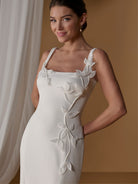A woman wearing a fitted white sleeveless dress with floral appliqué details along the bodice and side, posed against a warm tan studio background with draped white fabric at the side. / Square neckline scuba gown with handmade chiffon flowers.