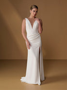A woman models a floor-length ivory wedding gown with a deep V-neckline, draped bodice, and thigh-high front slit, posed against a warm tan studio backdrop. / Slim gown with draped back and slit skirt.