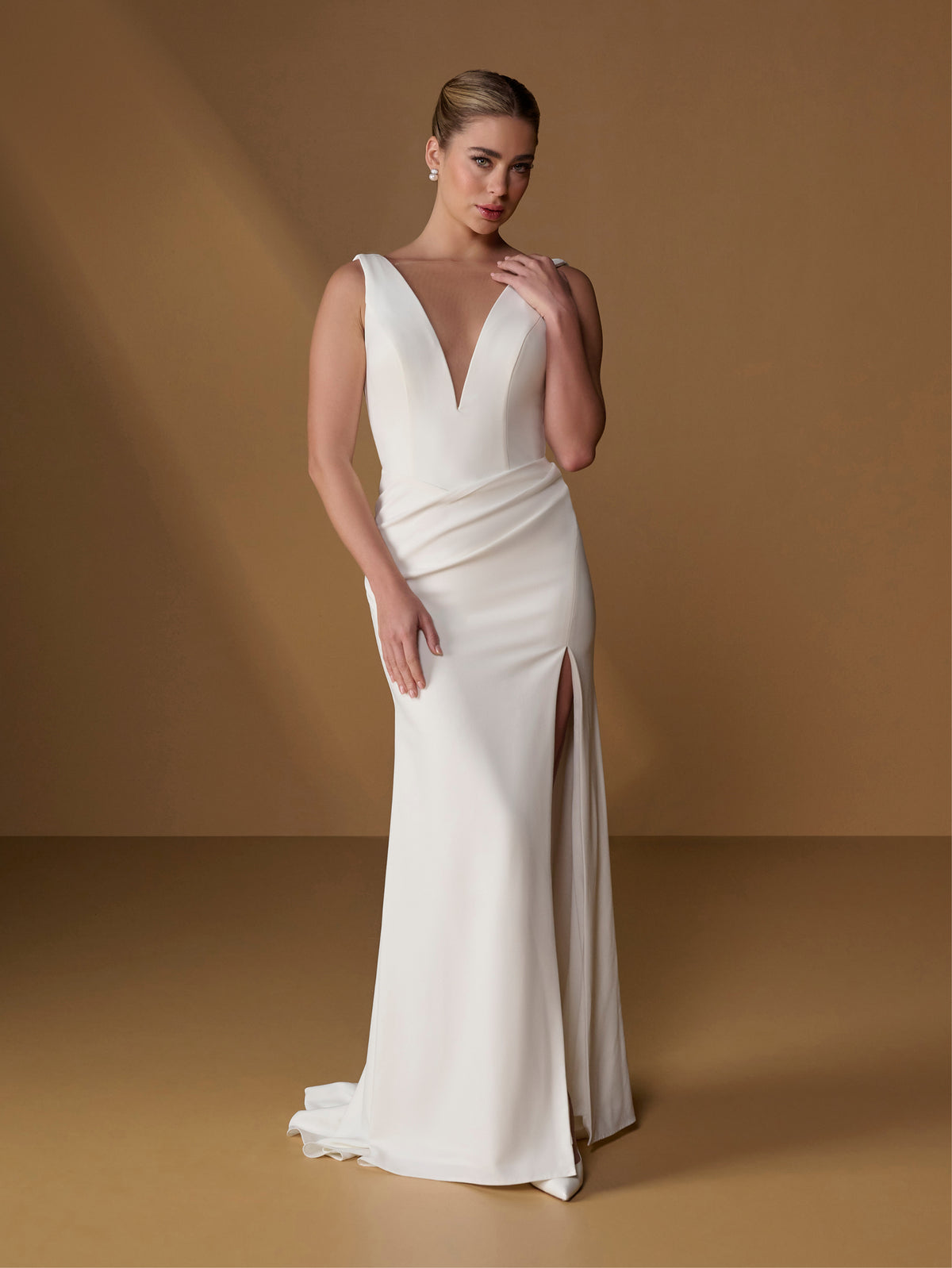 A woman models a floor-length ivory wedding gown with a deep V-neckline, draped bodice, and thigh-high front slit, posed against a warm tan studio backdrop. / Slim gown with draped back and slit skirt.