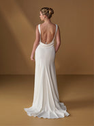 A woman modeling a floor-length ivory wedding gown from the back, showing a deep open back and draped train, set against a warm brown studio backdrop. / Slim gown with draped back and slit skirt.
