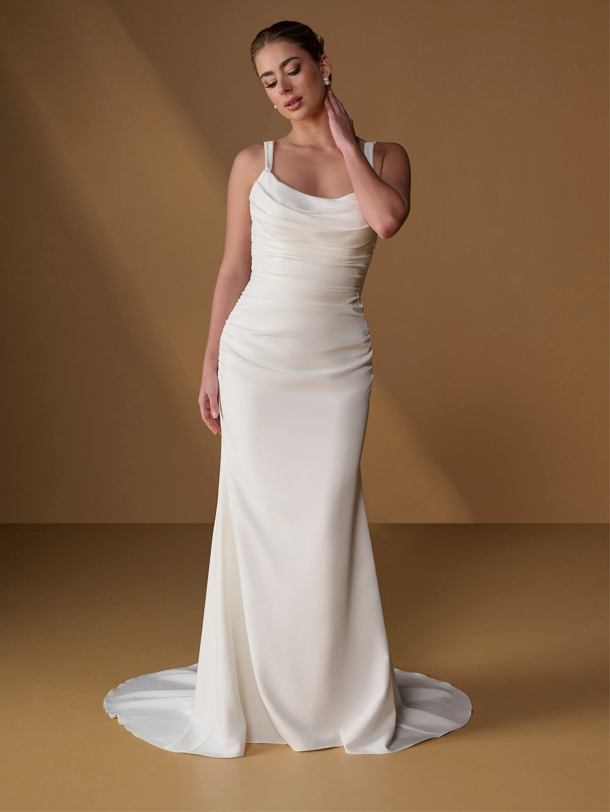 A woman modeling a floor-length ivory wedding dress with thin straps, a draped neckline, ruched bodice, and a slight train on a warm tan studio background. / Slim gown with shirring detail, cowl back and placed lace.