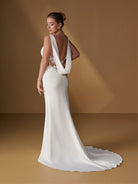 A woman modeling a white bridal gown from the back in a studio with a warm brown backdrop. The dress has a draped one-shoulder design, a deep open back, sheer lace detailing, and a long train. / Slim gown with shirring detail, cowl back and placed lace.