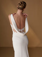 A woman shown from the back wearing a white bridal gown with a dramatic open back, sheer lace paneling, and wide draped straps, posed against a warm tan studio backdrop. / Slim gown with shirring detail, cowl back and placed lace.
