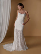 A woman models a sleeveless ivory lace wedding gown with thin straps and a fitted silhouette, standing in a warm tan studio setting with draped white fabric at the side. / Slim lace dress with short skirt lining. Scooped neckline with spaghetti straps. Zipper back closure.