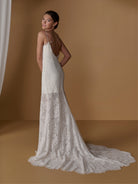 A woman modeling a sleeveless lace wedding gown with thin straps, a low open back, and a long train against a warm tan studio backdrop. / Slim lace dress with short skirt lining. Scooped neckline with spaghetti straps. Zipper back closure.
