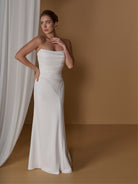 A woman models a strapless white floor-length gown with a fitted bodice and flowing skirt against a warm tan studio backdrop with draped white fabric on the left. / Slim gown with draping and asymmetrical seam detail.