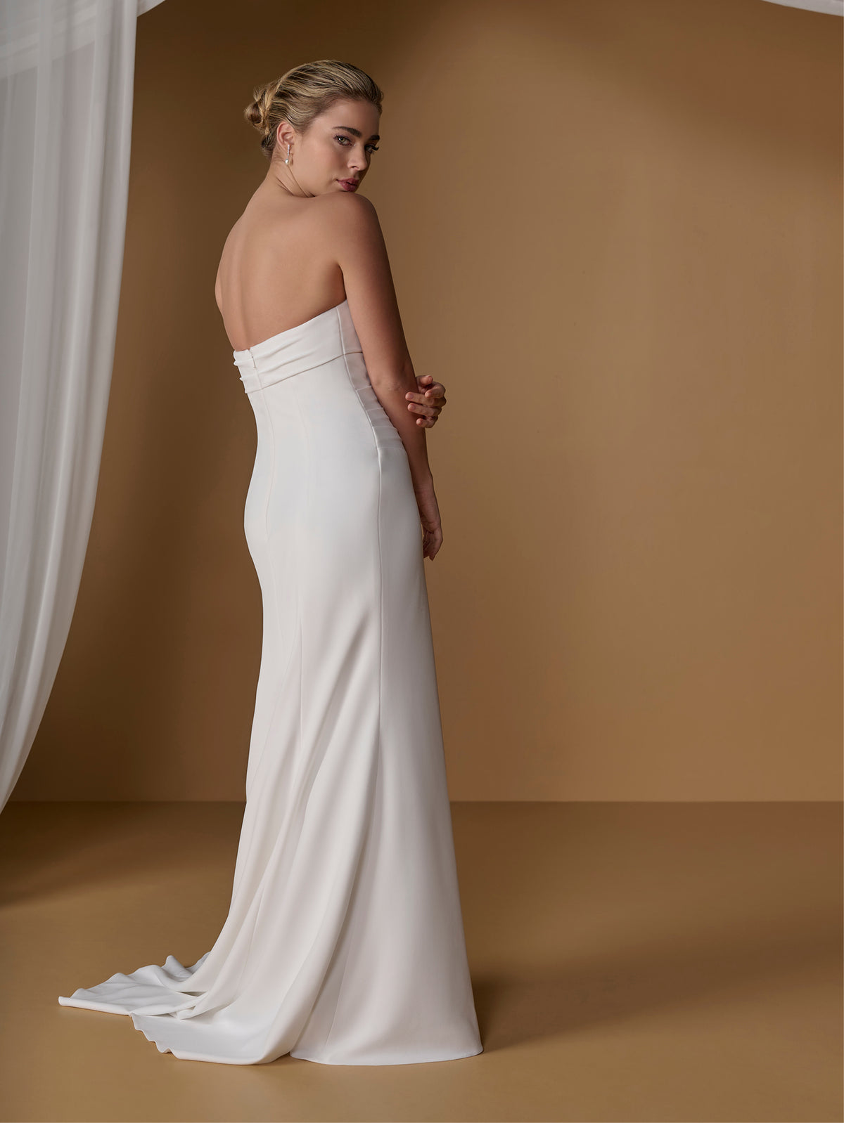 A woman modeling a strapless white wedding gown from the back, standing in a studio with a warm tan backdrop and a draped white fabric on the left. / Slim gown with draping and asymmetrical seam detail.