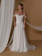 A woman models an off-the-shoulder A-line wedding gown with a pleated bodice and a long flowing train, posed against a warm tan studio backdrop with draped white fabric. / 2-piece crinkle chiffon gown with draped bodice and full skirt.