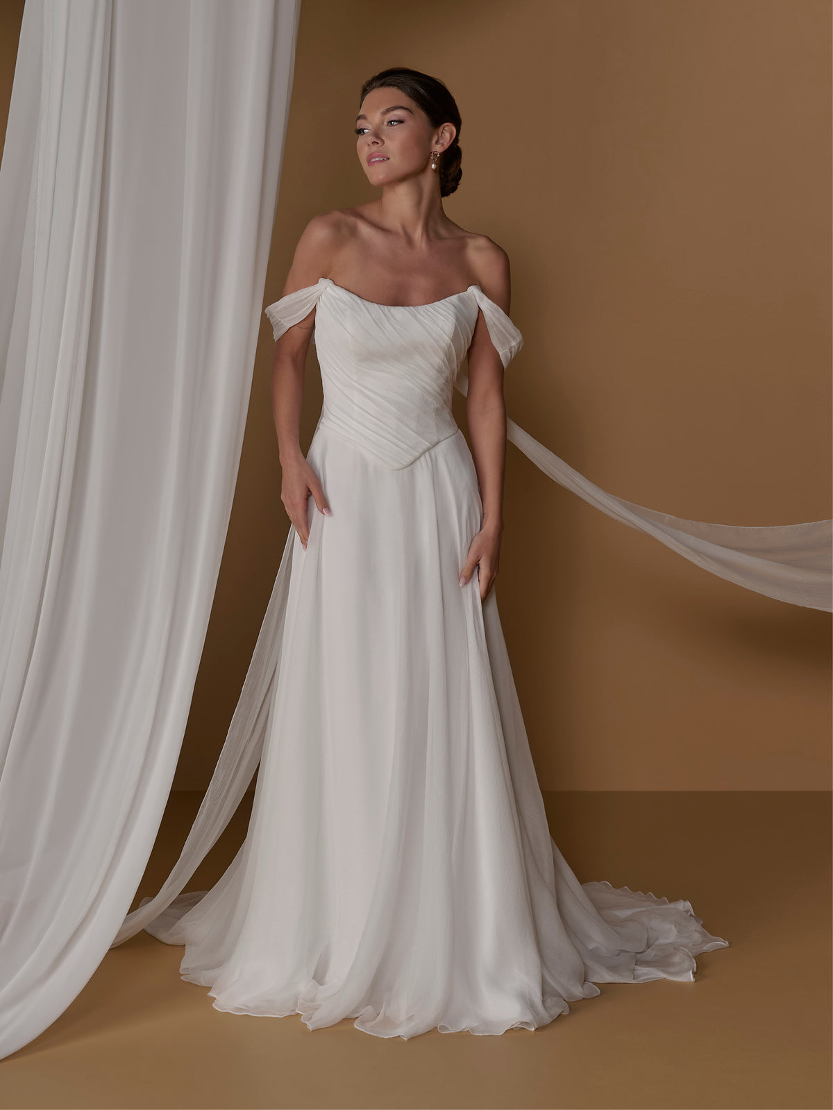 A woman models an off-the-shoulder A-line wedding gown with a pleated bodice and a long flowing train, posed against a warm tan studio backdrop with draped white fabric. / 2-piece crinkle chiffon gown with draped bodice and full skirt.