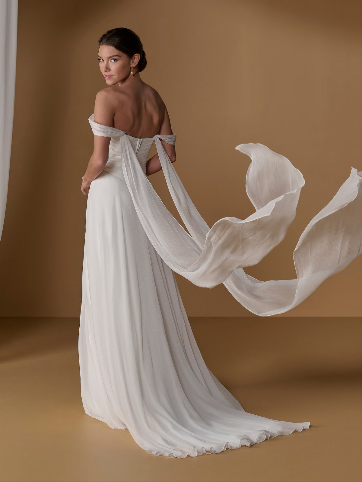 A woman modeling an off-the-shoulder white wedding gown with a long flowing train and sheer draped fabric, photographed against a warm tan studio backdrop. / 2-piece crinkle chiffon gown with draped bodice and full skirt.