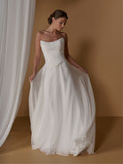 A woman modeling a strapless white wedding gown with a ruched bodice and full flowing skirt, posed against a warm tan studio backdrop with draped fabric on the left. / 2-piece crinkle chiffon gown with draped bodice and full skirt.