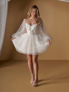 A woman modeling a short white bridal-style dress with a sweetheart neckline, sheer long balloon sleeves, a large bow at the bust, and a flared tulle skirt, posed against a warm tan studio backdrop. / Short dotted tulle gown with off the shoulder full sleeve and full skirt.