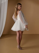 A woman modeling a short white tulle dress with sheer bubble sleeves, photographed in a warm tan studio setting with a white curtain on the left. / Short dotted tulle gown with off the shoulder full sleeve and full skirt.