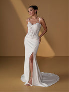 A woman models a white lace and satin wedding gown with thin straps, a fitted lace bodice, a high thigh slit, and a long train against a warm tan studio backdrop. / Lace bodice with lace up back, crepe skirt with side front split.