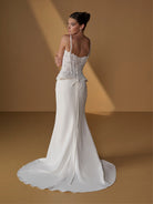 A woman modeling a white bridal gown from the back, standing on a warm tan studio floor with dramatic diagonal lighting. The dress has thin straps, a lace corset-style upper section, a fitted skirt, and a long train. / Lace bodice with lace up back, crepe skirt with side front split.