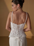 A woman shown from the back wearing a white lace corset-style top with thin straps and lace-up detailing, paired with a white skirt or dress, against a warm tan studio background. / Lace bodice with lace up back, crepe skirt with side front split.