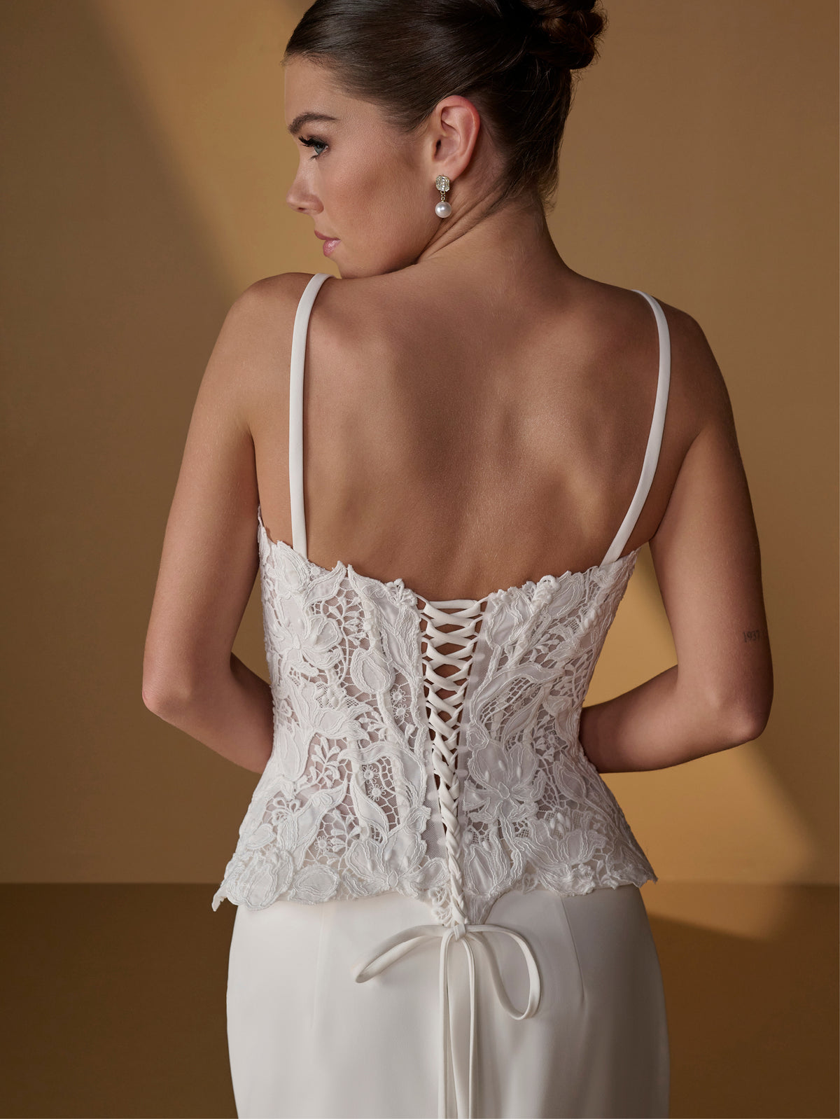 A woman shown from the back wearing a white lace corset-style top with thin straps and lace-up detailing, paired with a white skirt or dress, against a warm tan studio background. / Lace bodice with lace up back, crepe skirt with side front split.