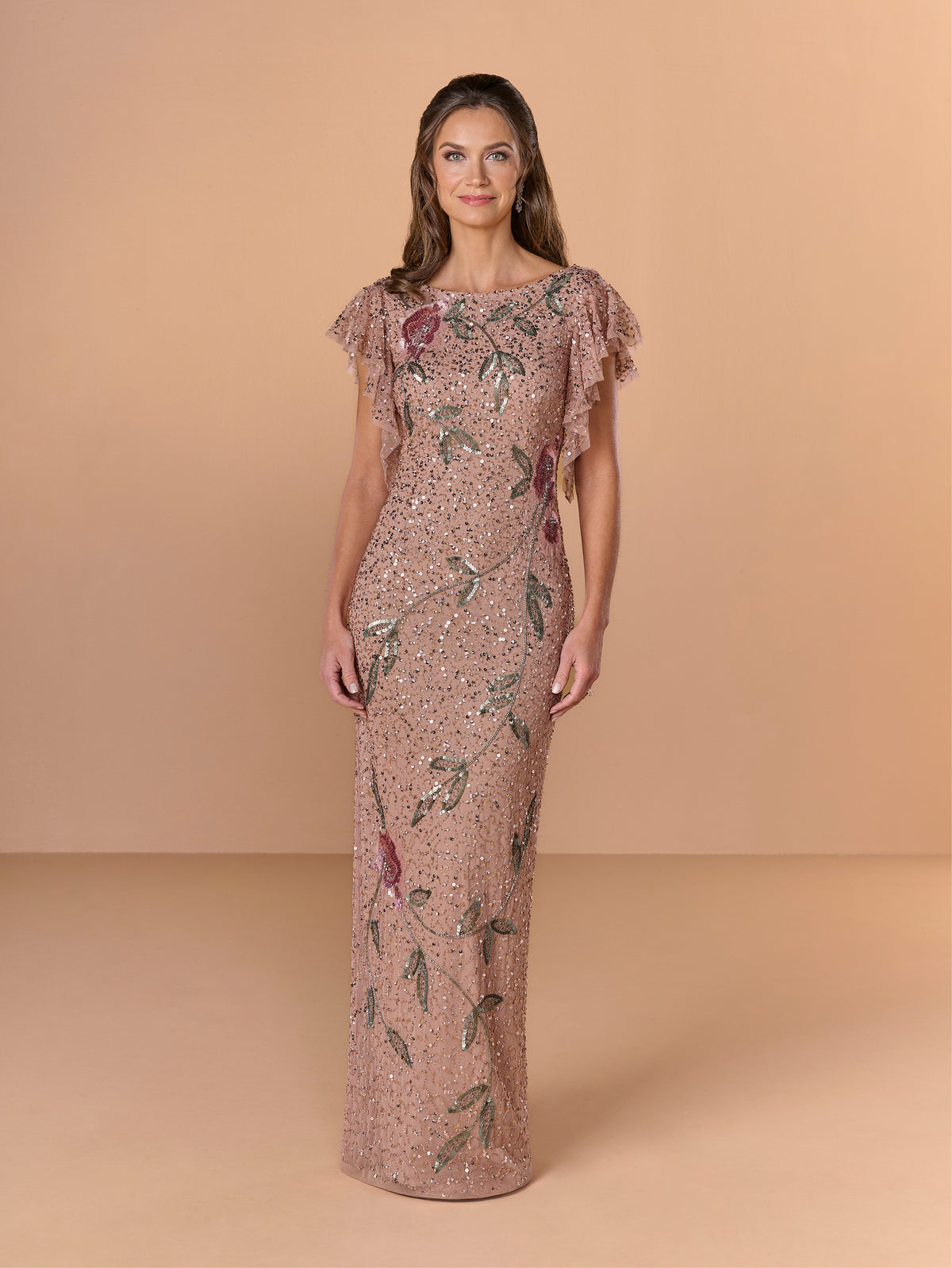 A woman modeling a floor-length, ruffled-sleeve evening gown in a dusty rose pink color with green and purple floral embroidery, set against a solid beige studio backdrop. / All over beaded column gown with flutter sleeve and back cowl.