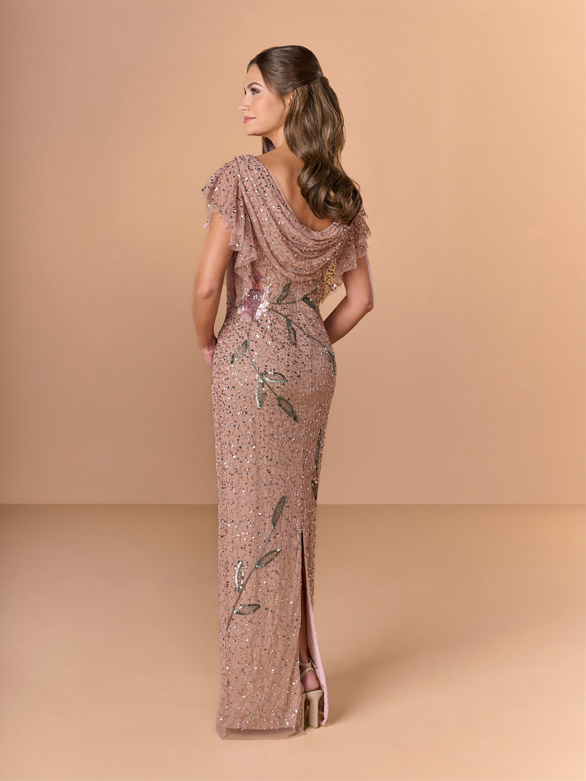 Woman wearing a sequin evening gown with floral embellishments on a beige background / All over beaded column gown with flutter sleeve and back cowl.