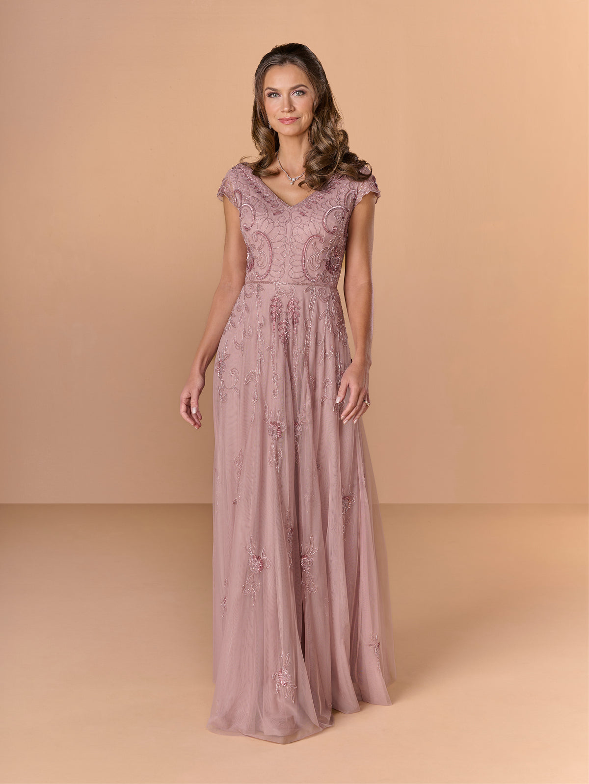Woman wearing a long, elegant dress with lace details against a beige background / All over beaded gown with sheer cap sleeve, v neckline and full, A-line skirt.